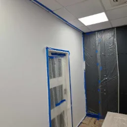 Przygotowanie pomieszczenia do malowania: białe ściany zabezpieczone niebieską taśmą malarską, drzwi i szafa w folii ochronnej, podłoga wyłożona kartonem.