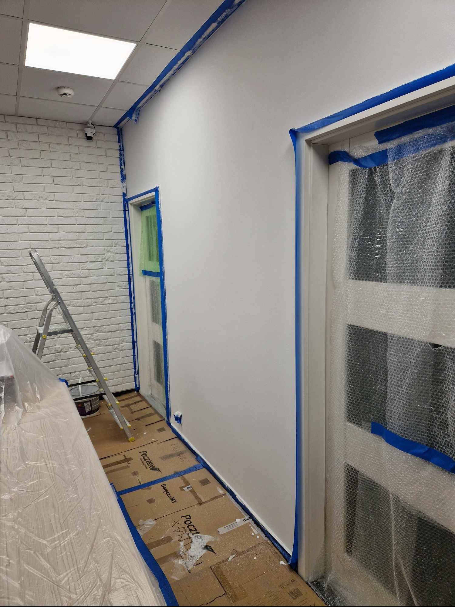 Przygotowanie do malowania wnętrza: ściany i framugi zabezpieczone taśmą malarską, podłoga wyłożona kartonami, drzwi osłonięte folią bąbelkową, drabina oparta o ścianę z cegły dekoracyjnej.