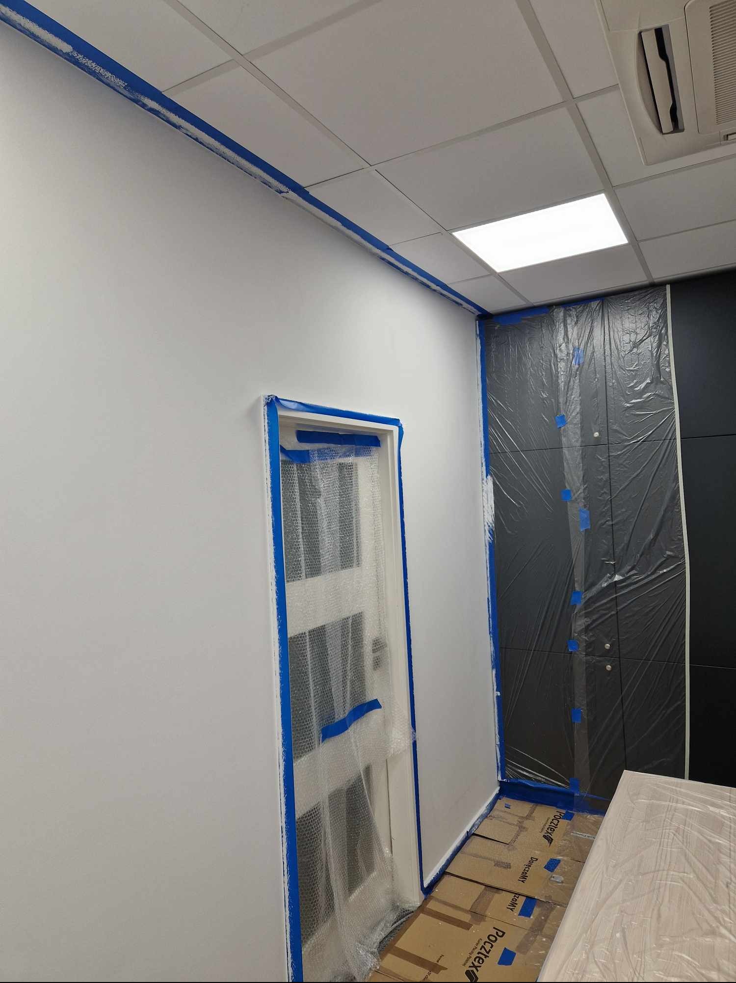 Przygotowanie pomieszczenia do malowania: białe ściany zabezpieczone niebieską taśmą malarską, drzwi i szafa w folii ochronnej, podłoga wyłożona kartonem.