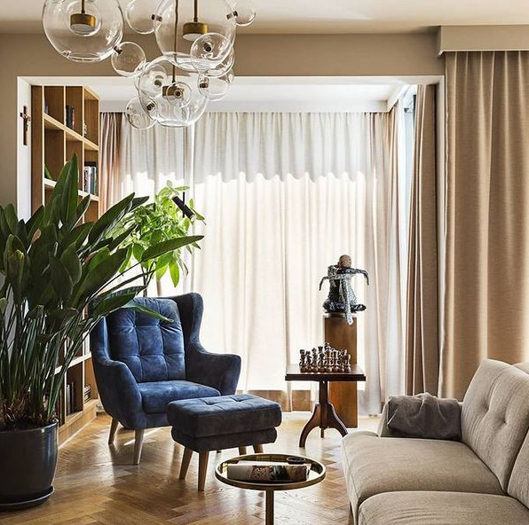 Elegancki salon z parkietem w jodełkę, głębokim niebieskim fotelem typu uszak z podnóżkiem, beżową sofą, szklanym żyrandolem w kształcie bąbelków i szachami na stoliku