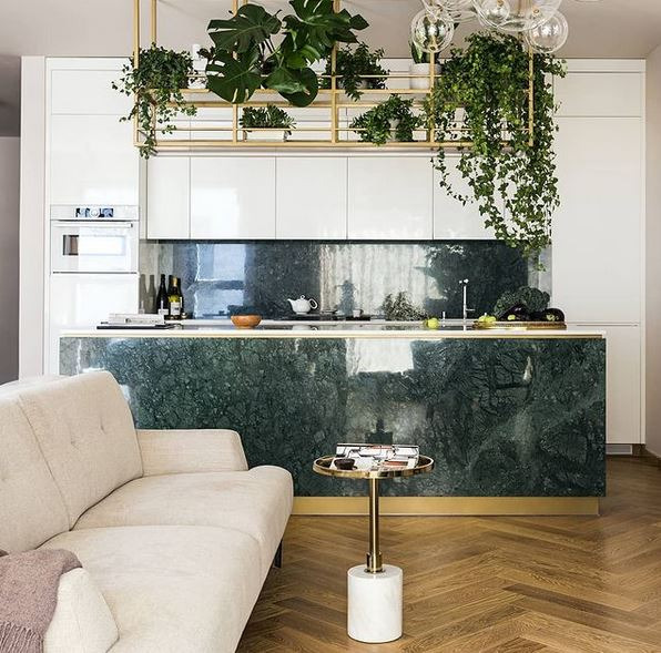 Wnętrze kuchni z wyspą wykończoną zielonym marmurem, nad którą zwisa metalowa konstrukcja z roślinami doniczkowymi. W tle białe szafki i sprzęty AGD, a z boku fragment kremowej sofy i stolika.