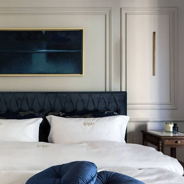 Elegancka sypialnia z łóżkiem o granatowym, pikowanym wezgłowiu, białymi poduszkami z haftowanymi inicjałami i obrazem w złotej ramie. Po prawej stronie minimalistyczna lampa wisząca i stolik nocny...