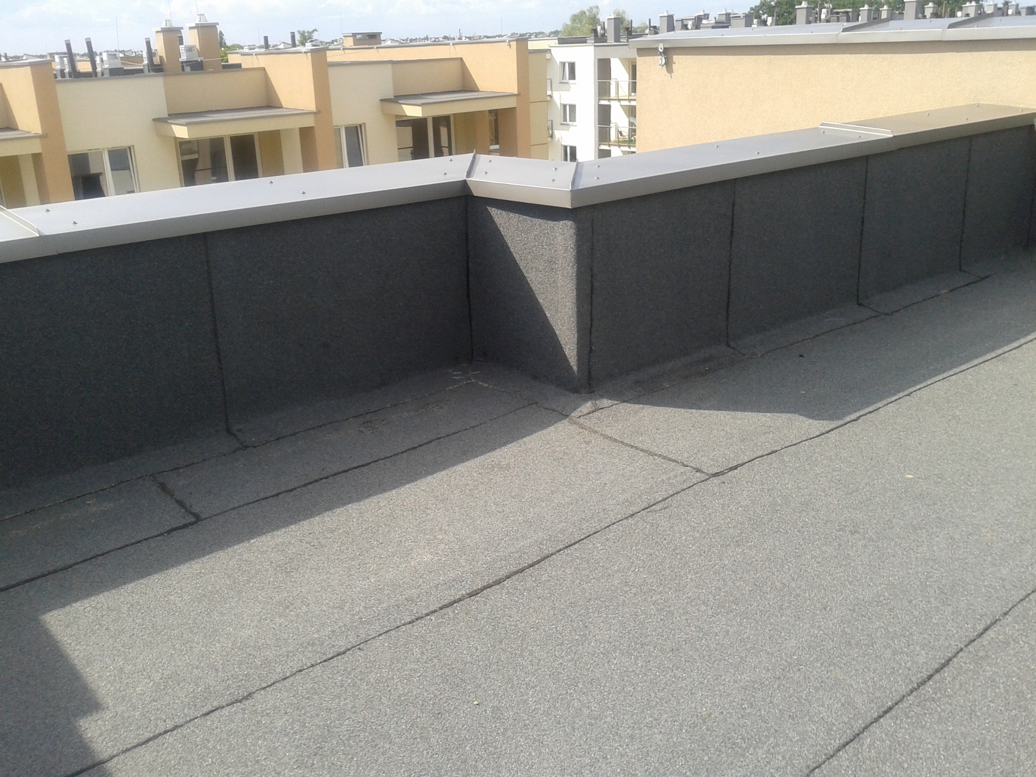 Płaski dach pokryty papą termozgrzewalną z obróbką blacharską na krawędzi, widok z góry na tle budynków mieszkalnych.