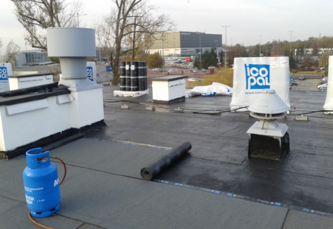 Niedawno położona, czarna papa na płaskim dachu, widoczna butla gazowa, rolka papy, kominki wentylacyjne i opakowania materiałów budowlanych na tle parkingu i budynku.