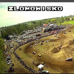 Szeroki widok z lotu ptaka na teren złomowiska z torem wyścigowym, gdzie odbywają się wyścigi samochodowe, otoczony drzewami i budynkami.