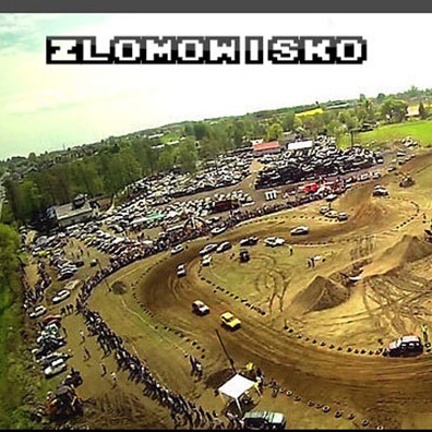 Szeroki widok z lotu ptaka na teren złomowiska z torem wyścigowym, gdzie odbywają się wyścigi samochodowe, otoczony drzewami i budynkami.