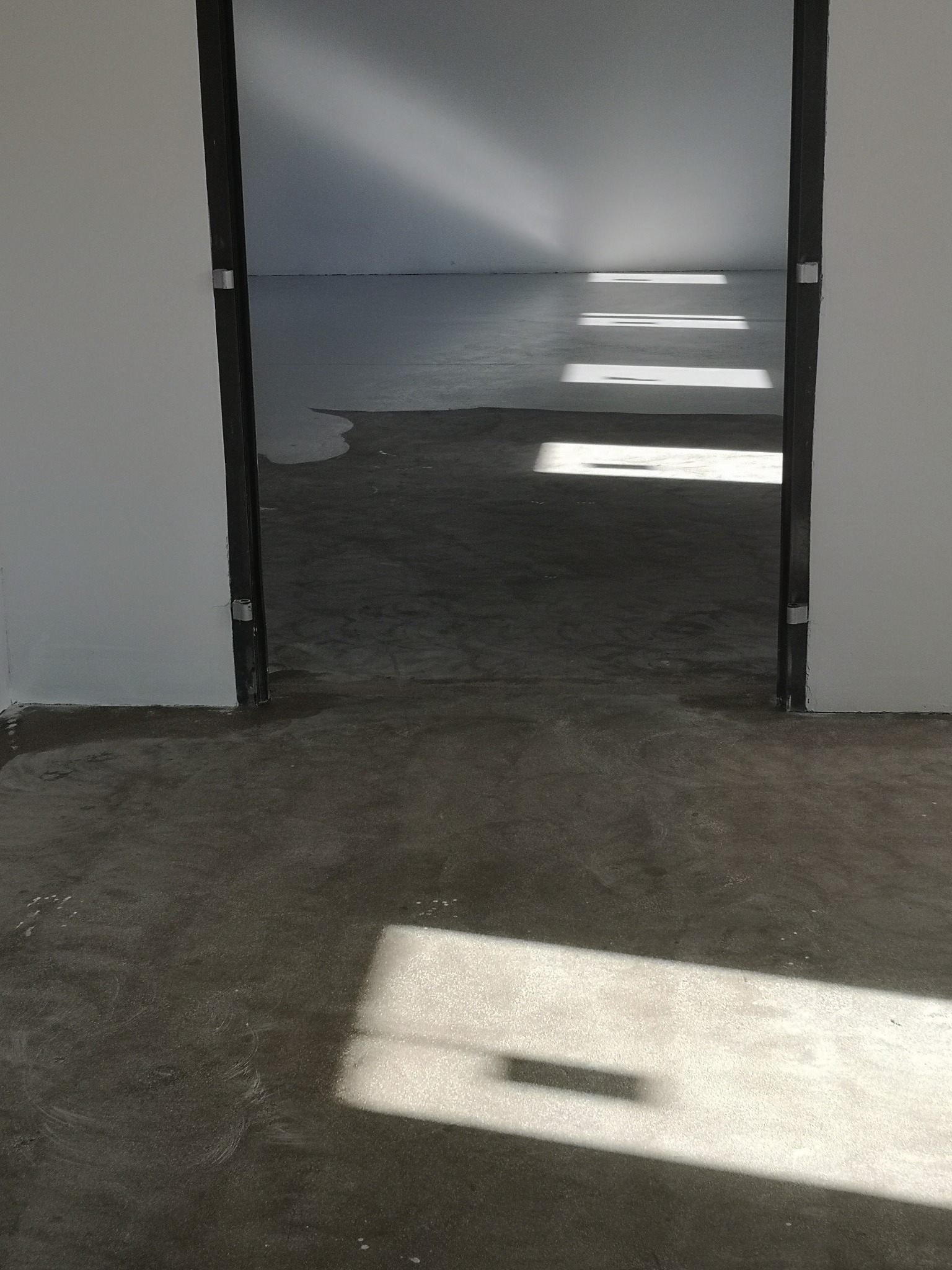 Szara, wylewana podłoga w surowym stanie, widziana przez framugę drzwi, z widocznymi smugami i refleksami światła słonecznego wpadającego przez okno.
