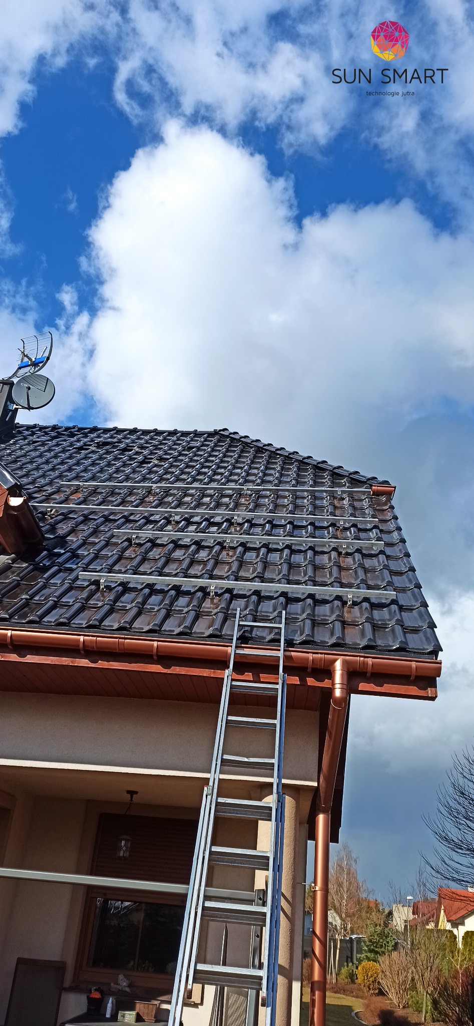 Montaż szyn pod panele fotowoltaiczne na błyszczącym, brązowym dachu z dachówki ceramicznej, drabina oparta o dach, na tle błękitnego nieba z obłokami.