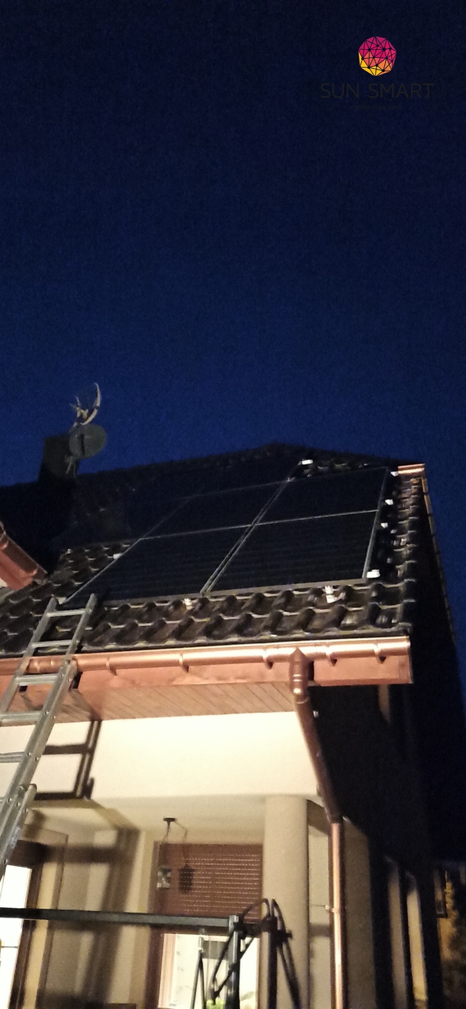 Panele słoneczne na dachu domu z miedzianymi rynnami, drabina oparta o dach, logo firmy widoczne w górnym rogu, nocne ujęcie.