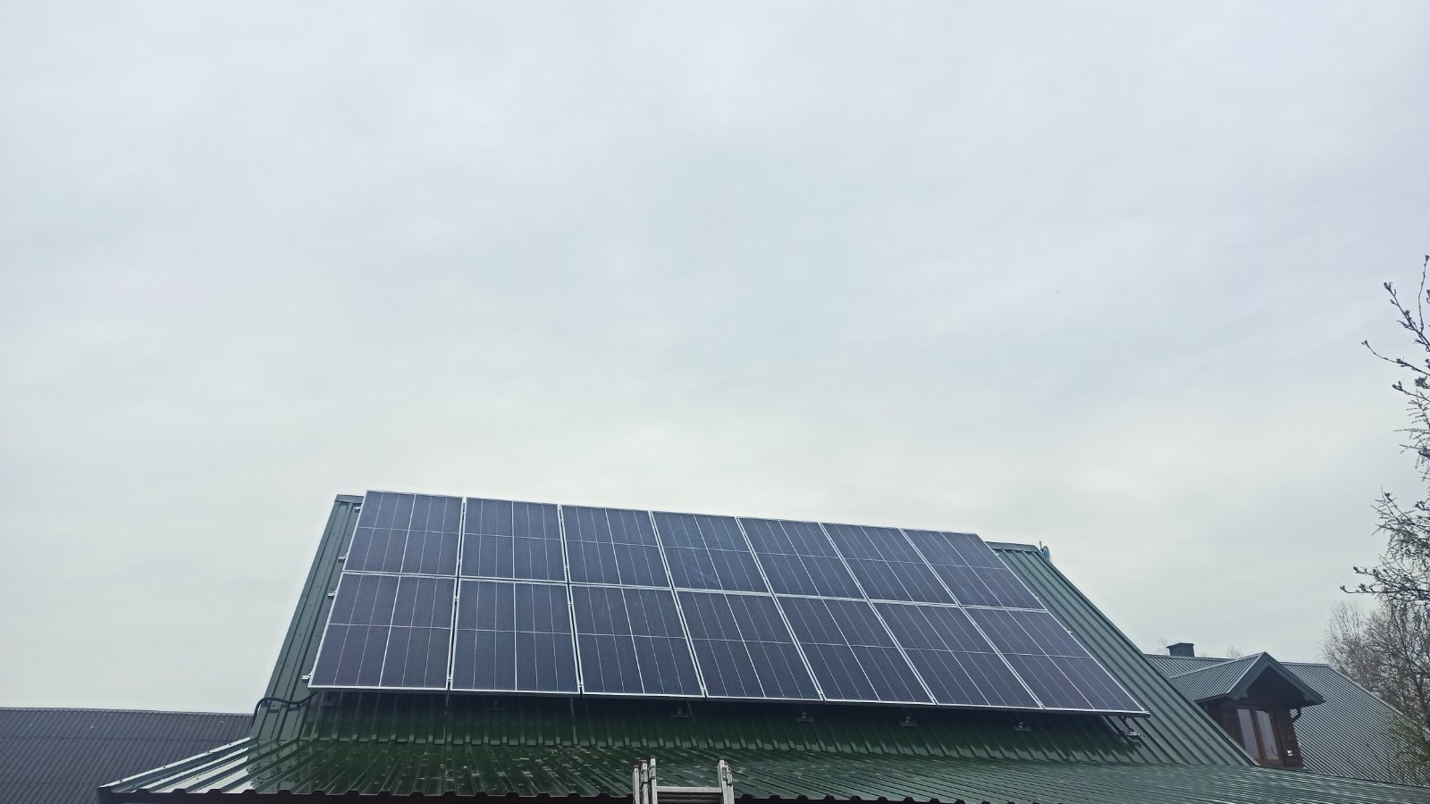 Panel słoneczny na zielonym, metalowym dachu domu w Biłgoraju, widoczne drabiny opierające się o dach, pochmurne niebo w tle.