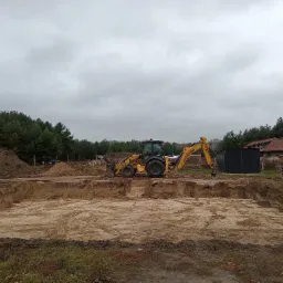 Żółta koparko-ładowarka New Holland na skraju wykopu pod fundamenty domu jednorodzinnego, widoczny teren budowy z usypanym piaskiem, w tle las i pochmurne niebo.