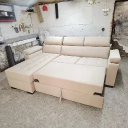 Rozłożona, jasnobeżowa sofa z wysuwanym siedziskiem i schowkiem, widoczna w warsztacie z narzędziami na ścianie i surowym betonem.