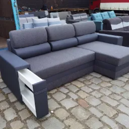 Szara, rozkładana sofa narożna z wysuwaną szufladą w podłokietniku, na tle kostki brukowej i innych mebli w magazynie.