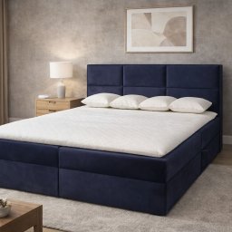 Tapicerowane, granatowe łóżko kontynentalne z wezgłowiem w kwadraty, trzy poduszki i biały materac. Na stoliku nocnym lampka. W tle szara ściana i obraz.