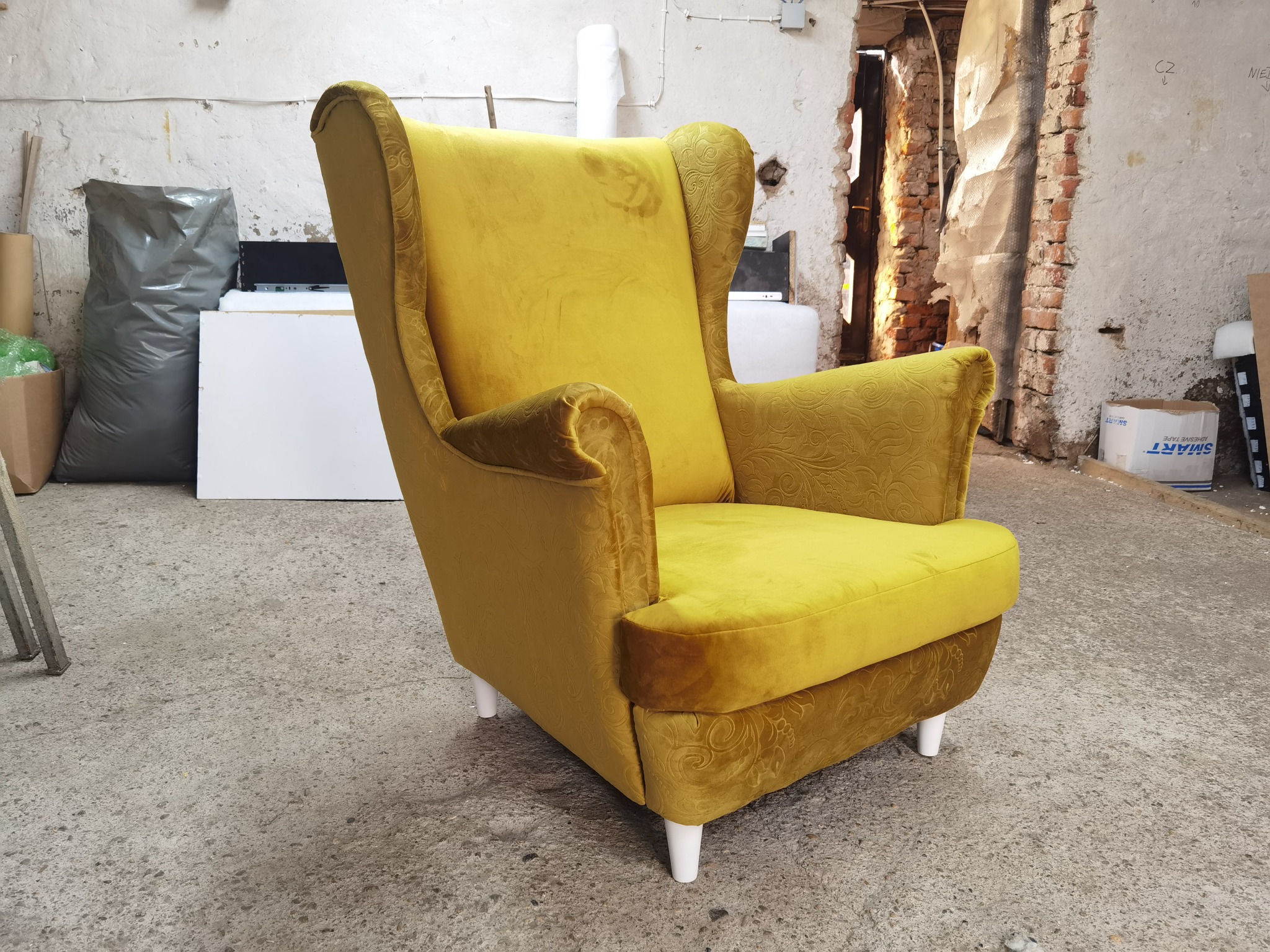 Żółty fotel uszak z weluru w warsztacie, widoczne białe nóżki i wzór na bokach, surowe tło z cegłą i widocznymi śladami remontu.