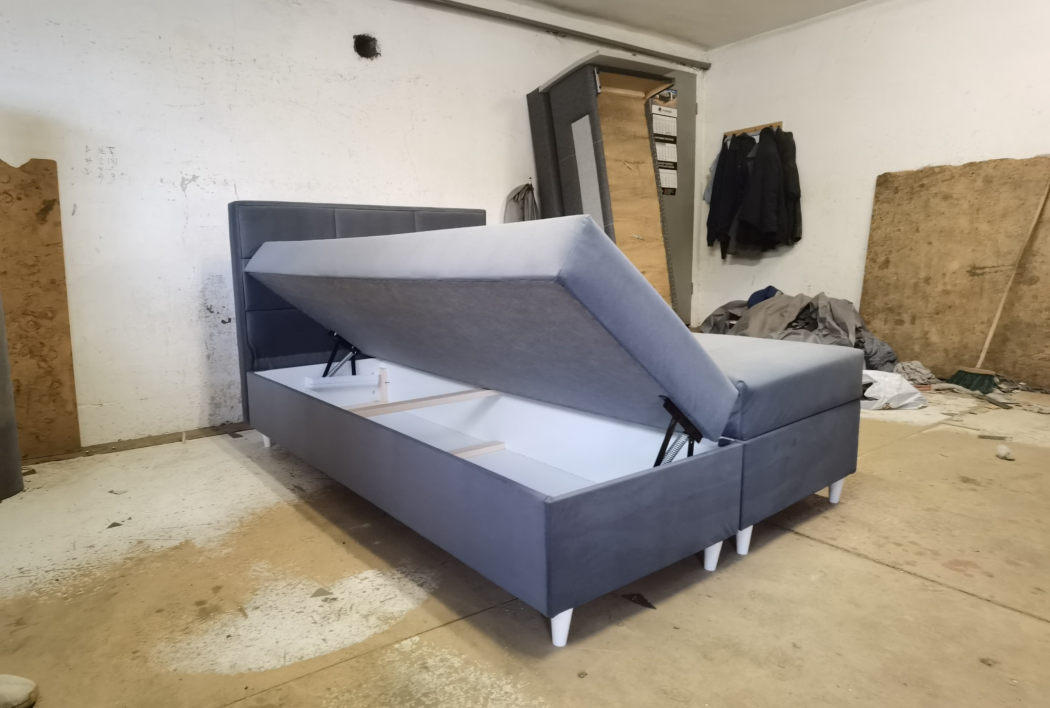 Szara tapicerowana sofa z podnoszonym siedziskiem, ukazująca biały pojemnik na pościel wewnątrz, na tle surowego wnętrza warsztatu.