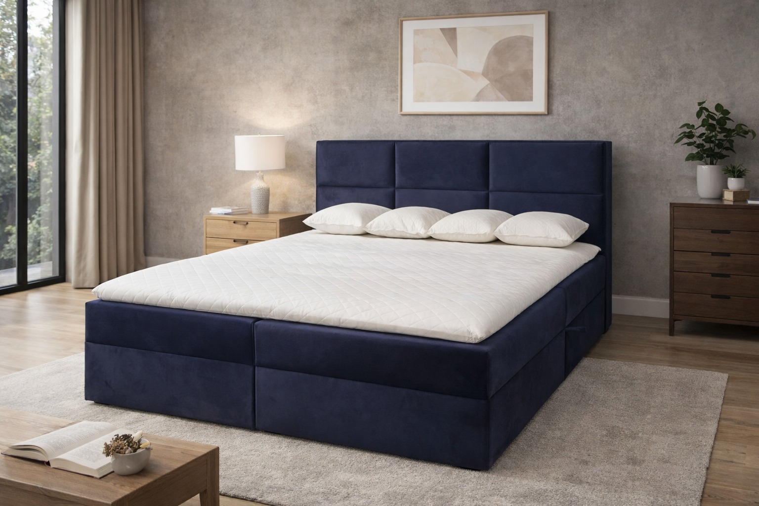 Tapicerowane, granatowe łóżko kontynentalne z wezgłowiem w kwadraty, trzy poduszki i biały materac. Na stoliku nocnym lampka. W tle szara ściana i obraz.