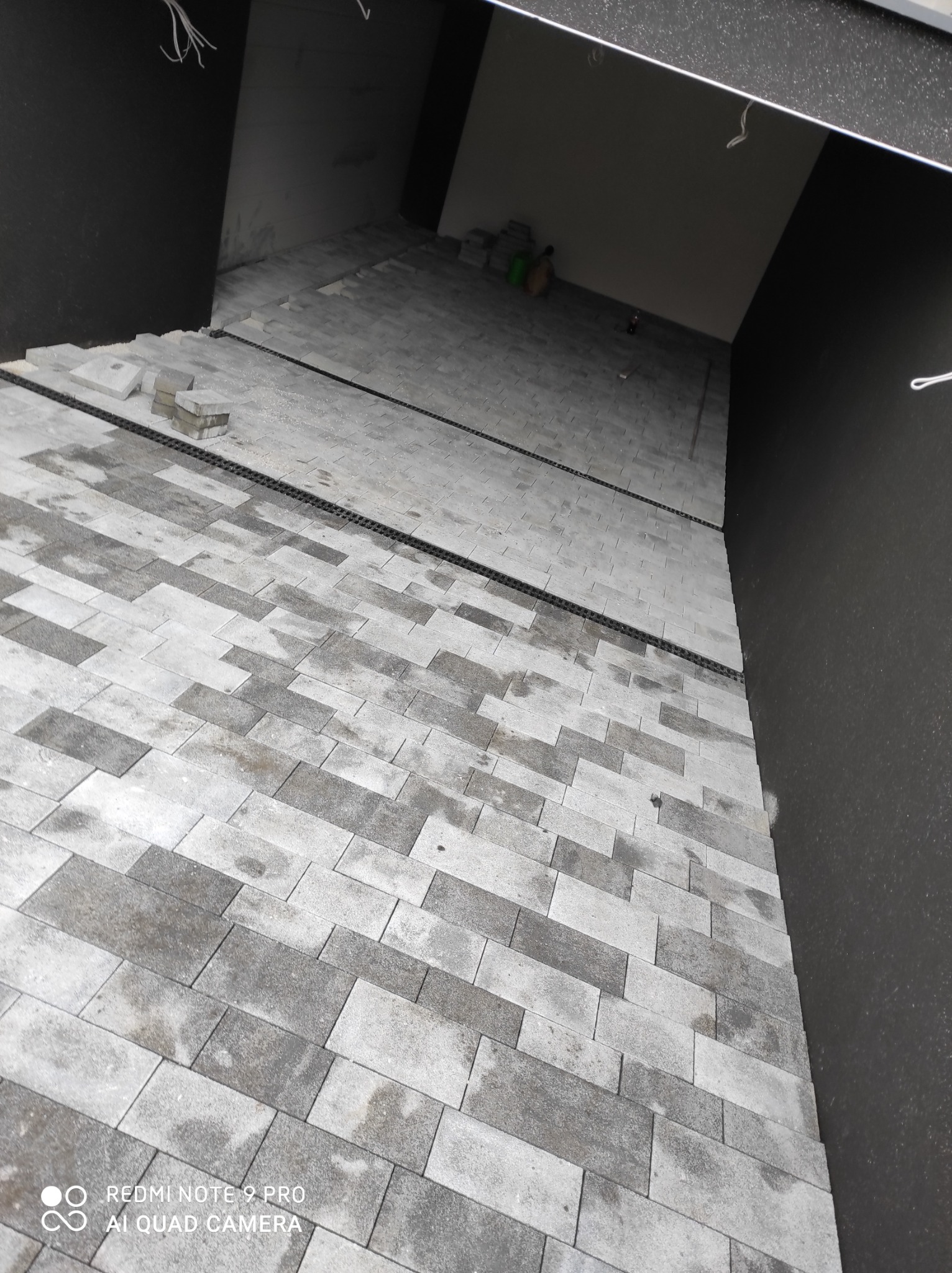 Ukończona nawierzchnia z szarej kostki brukowej ułożonej we wzór cegiełki, z widocznym systemem odprowadzania wody w postaci czarnych korytek wzdłuż krawędzi, częściowo pokryta wilgocią.