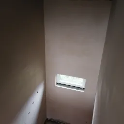 Świeżo otynkowane ściany wewnętrzne z małym oknem, widoczne otwory po kołkach na ścianie po lewej stronie, surowy strop z drewnianymi belkami, betonowa podłoga w trakcie prac wykończeniowych.