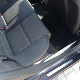 Wnętrze samochodu: tylna kanapa z szarej tapicerki, czarna wykładzina podłogowa i widoczny próg. Widoczne są elementy mocowania pasów bezpieczeństwa. Zdjęcie zrobione z bliska, z perspektywy dolnej.