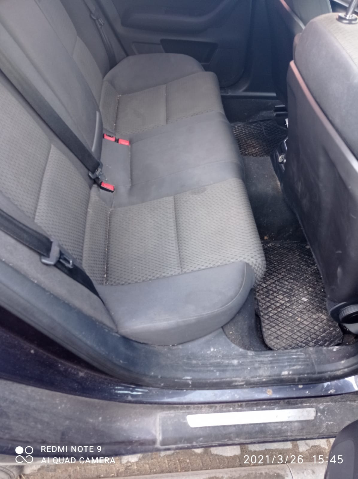 Tylna kanapa samochodu przed czyszczeniem, widoczne zabrudzenia na tapicerce i dywanikach, pasy bezpieczeństwa z czerwonymi elementami, ciemna kolorystyka wnętrza.