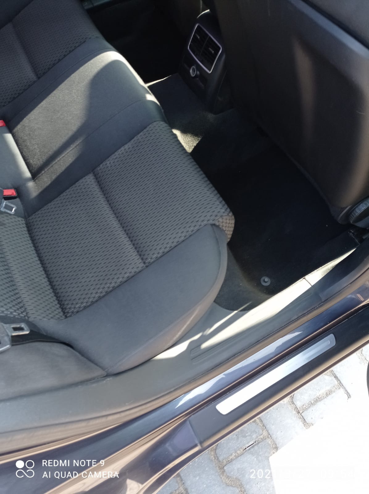 Wnętrze samochodu: tylna kanapa z szarej tapicerki, czarna wykładzina podłogowa i widoczny próg. Widoczne są elementy mocowania pasów bezpieczeństwa. Zdjęcie zrobione z bliska, z perspektywy dolnej.