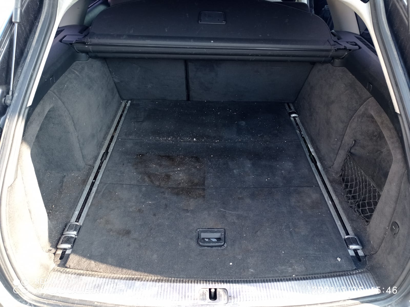 Brudny bagażnik samochodu z plamami i okruchami, widoczne metalowe szyny i siatka po prawej stronie.