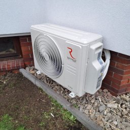Green Energy Łukasz Wieczorek - Biała jednostka zewnętrzna klimatyzacji Rotenso Hiro, zamontowana na szarym podłożu z kamieni, przy ceglanym cokole budynku z białą elewacją.