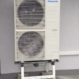 Biała jednostka zewnętrzna klimatyzacji marki Panasonic, zamontowana na stalowej konstrukcji wsporczej z betonowymi podstawami, na tle szarej ściany budynku.