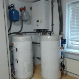Dwa białe, cylindryczne zbiorniki owinięte folią ochronną, zainstalowane w pomieszczeniu z białymi ścianami i brązową podłogą, widoczne rury i zawory, na ścianie zamontowany biały piec grzewczy...
