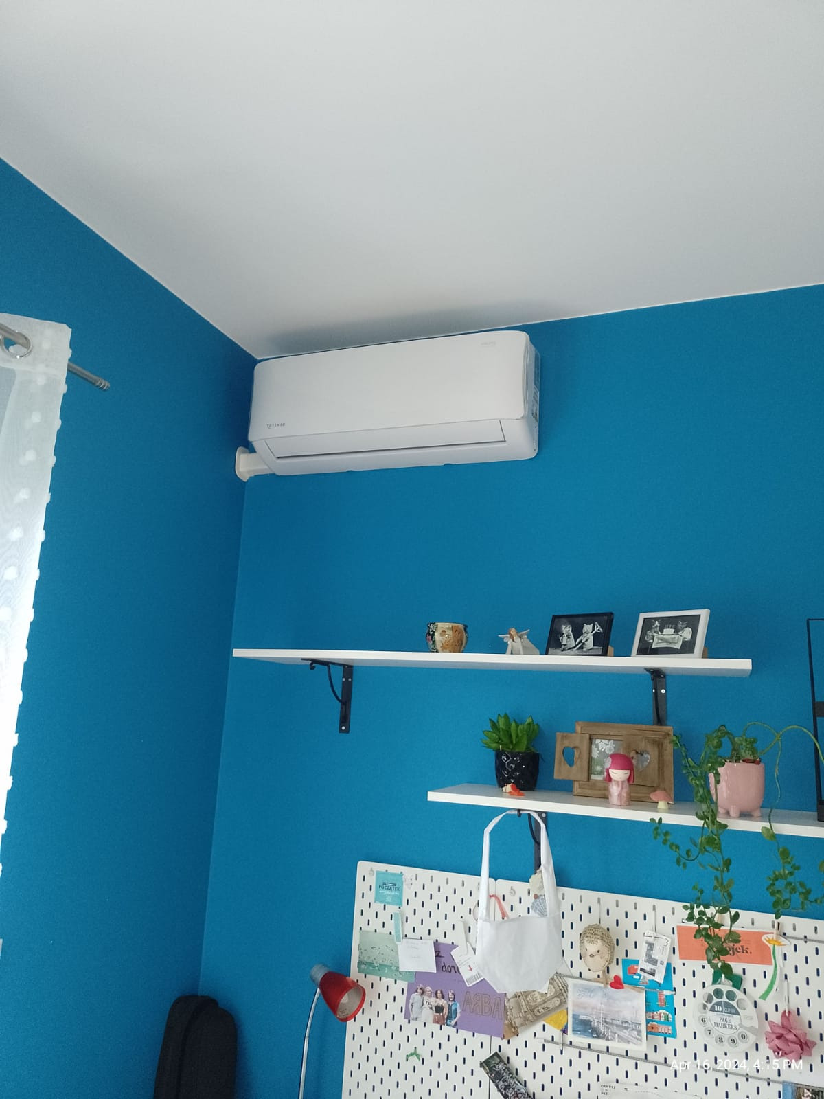 Biały klimatyzator na błękitnej ścianie w pokoju, widoczne białe półki z dekoracjami i tablica korkowa z notatkami.