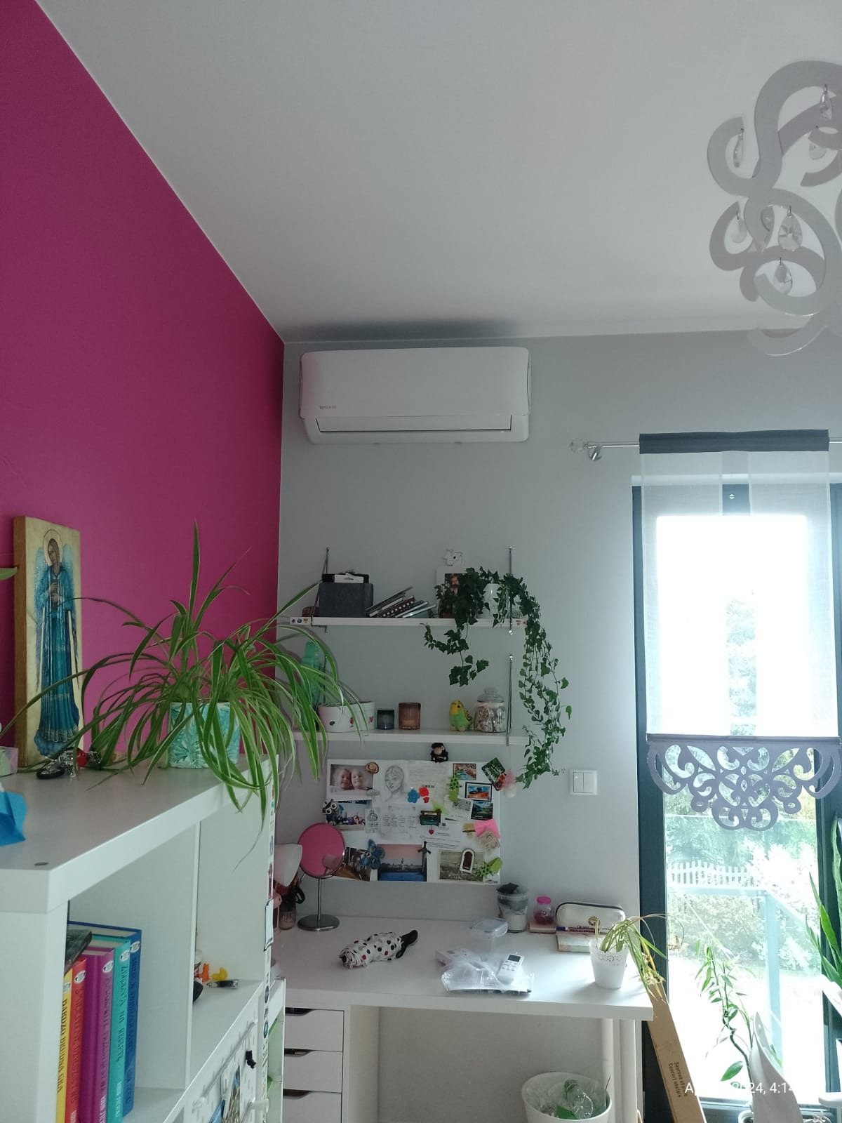 Biały klimatyzator ścienny zamontowany w pokoju nad biurkiem, widok na ścianę pomalowaną częściowo na różowo, na biurku rośliny w doniczkach i inne przedmioty.