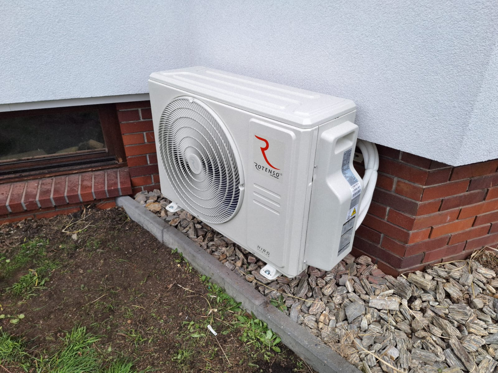 Biała jednostka zewnętrzna klimatyzacji Rotenso Hiro, zamontowana na szarym podłożu z kamieni, przy ceglanym cokole budynku z białą elewacją.