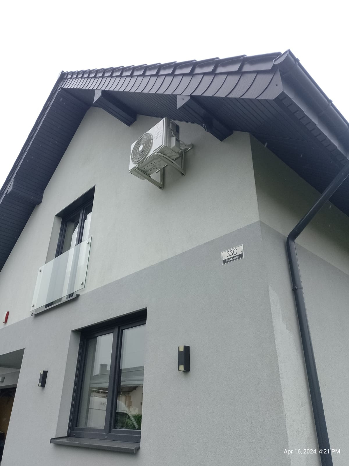 Zewnętrzna jednostka klimatyzacji zamontowana na elewacji dwukondygnacyjnego domu z szarą elewacją, widoczny numer domu 33C, czarne okna i balkon z szklaną balustradą.