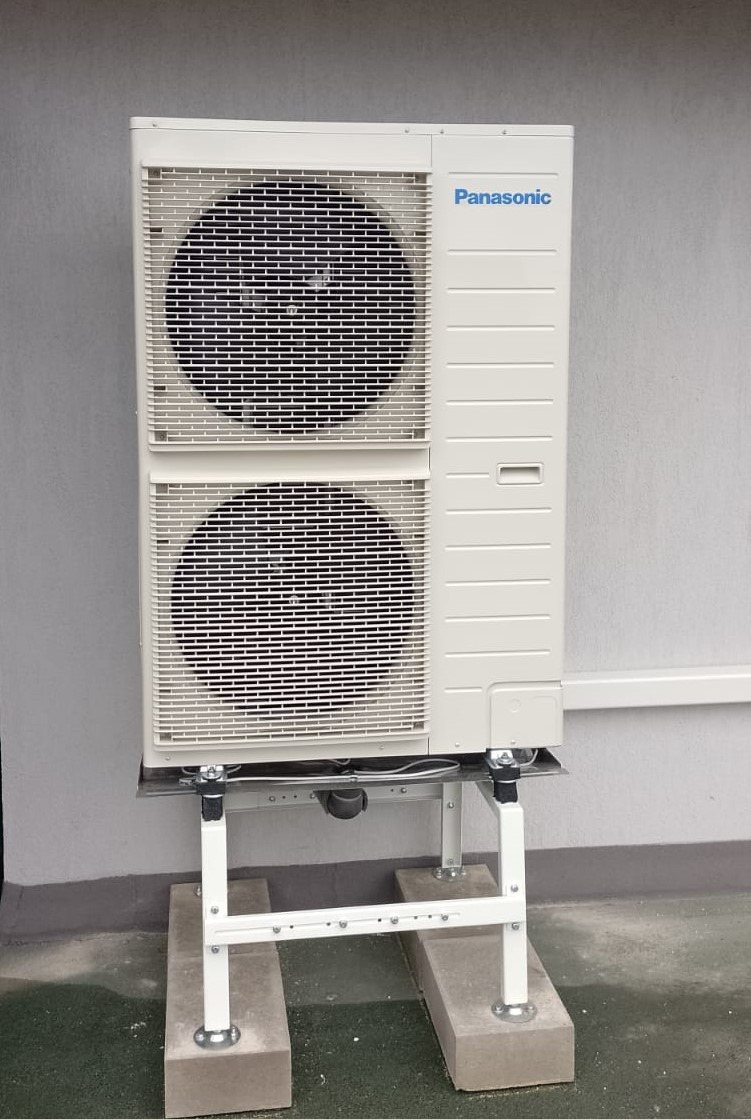 Biała jednostka zewnętrzna klimatyzacji marki Panasonic, zamontowana na stalowej konstrukcji wsporczej z betonowymi podstawami, na tle szarej ściany budynku.