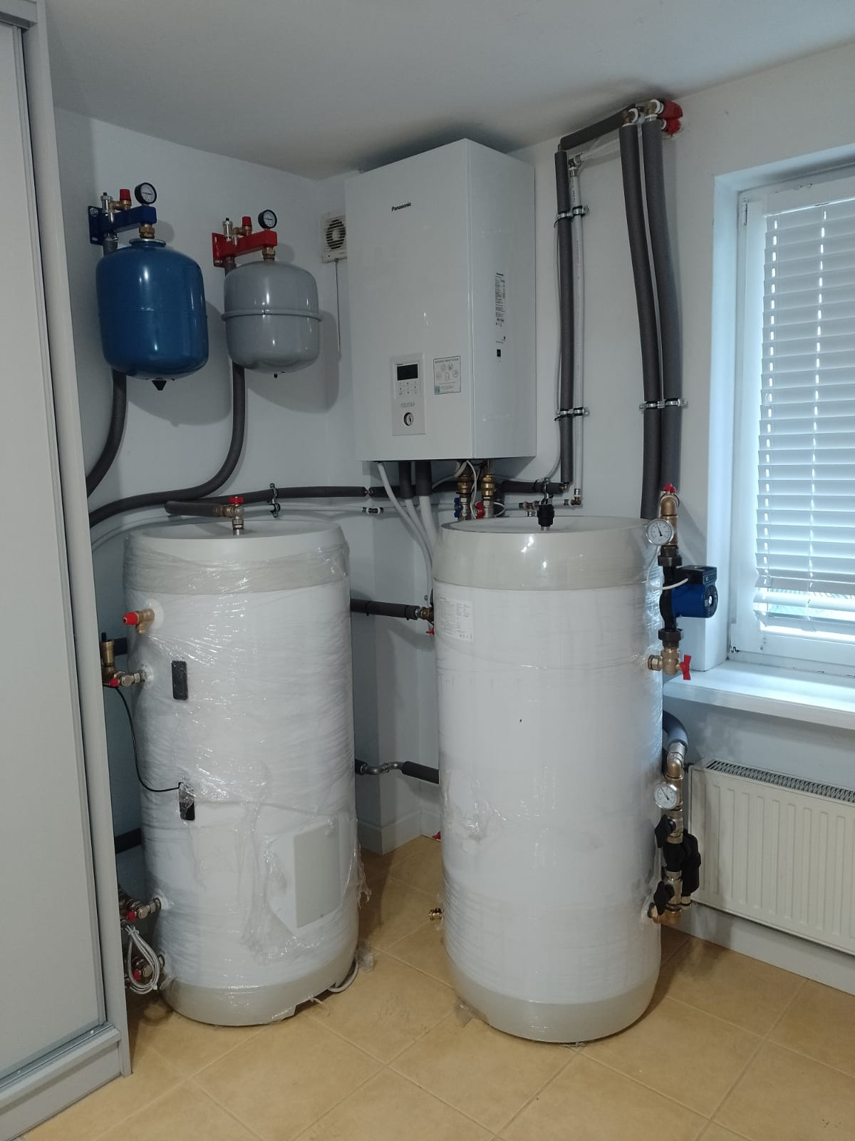 Dwa białe, cylindryczne zbiorniki owinięte folią ochronną, zainstalowane w pomieszczeniu z białymi ścianami i brązową podłogą, widoczne rury i zawory, na ścianie zamontowany biały piec grzewczy...
