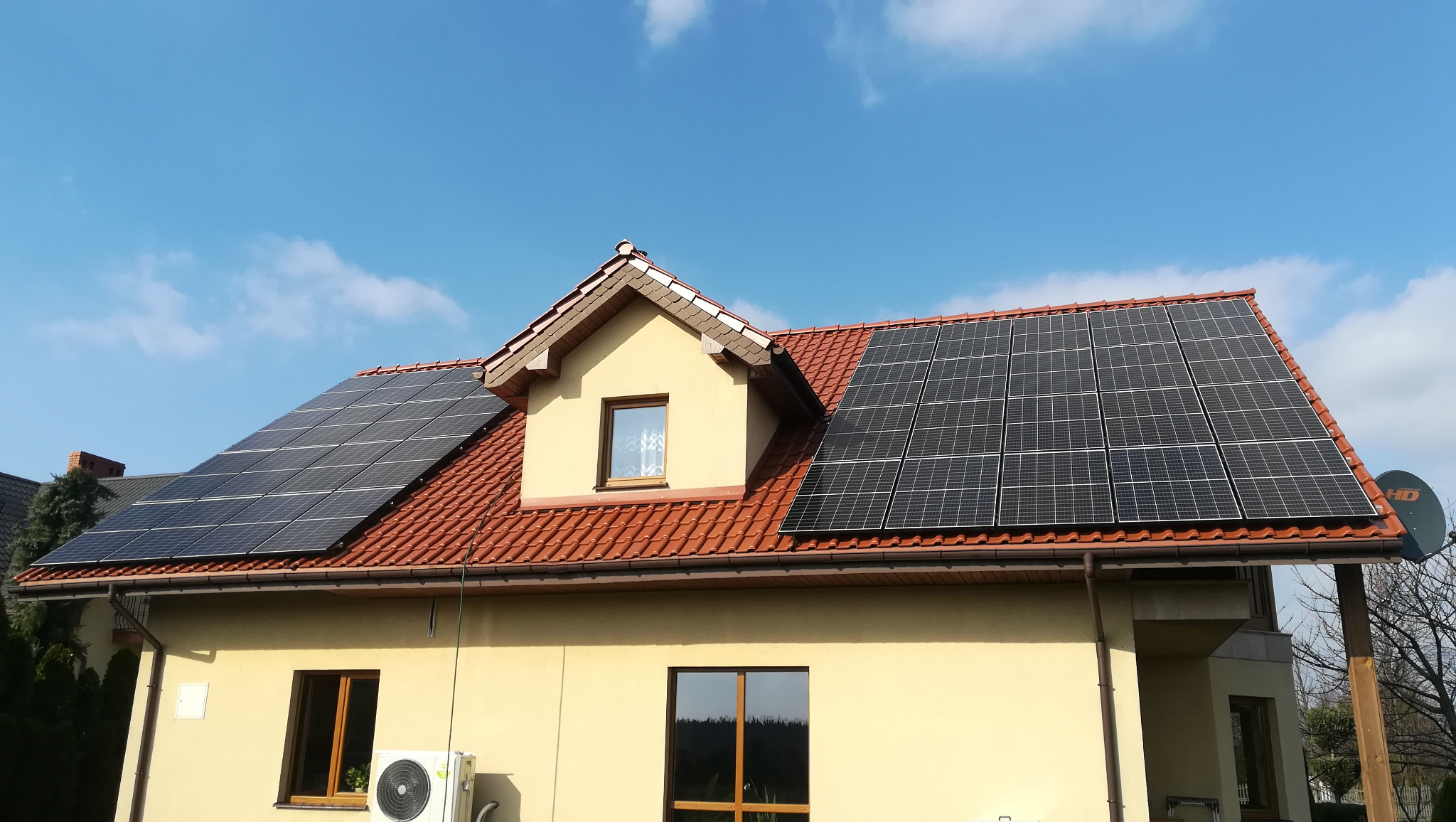 Dom jednorodzinny z panelami słonecznymi na dachu z czerwonej dachówki, widoczna jednostka klimatyzacji na ścianie budynku.