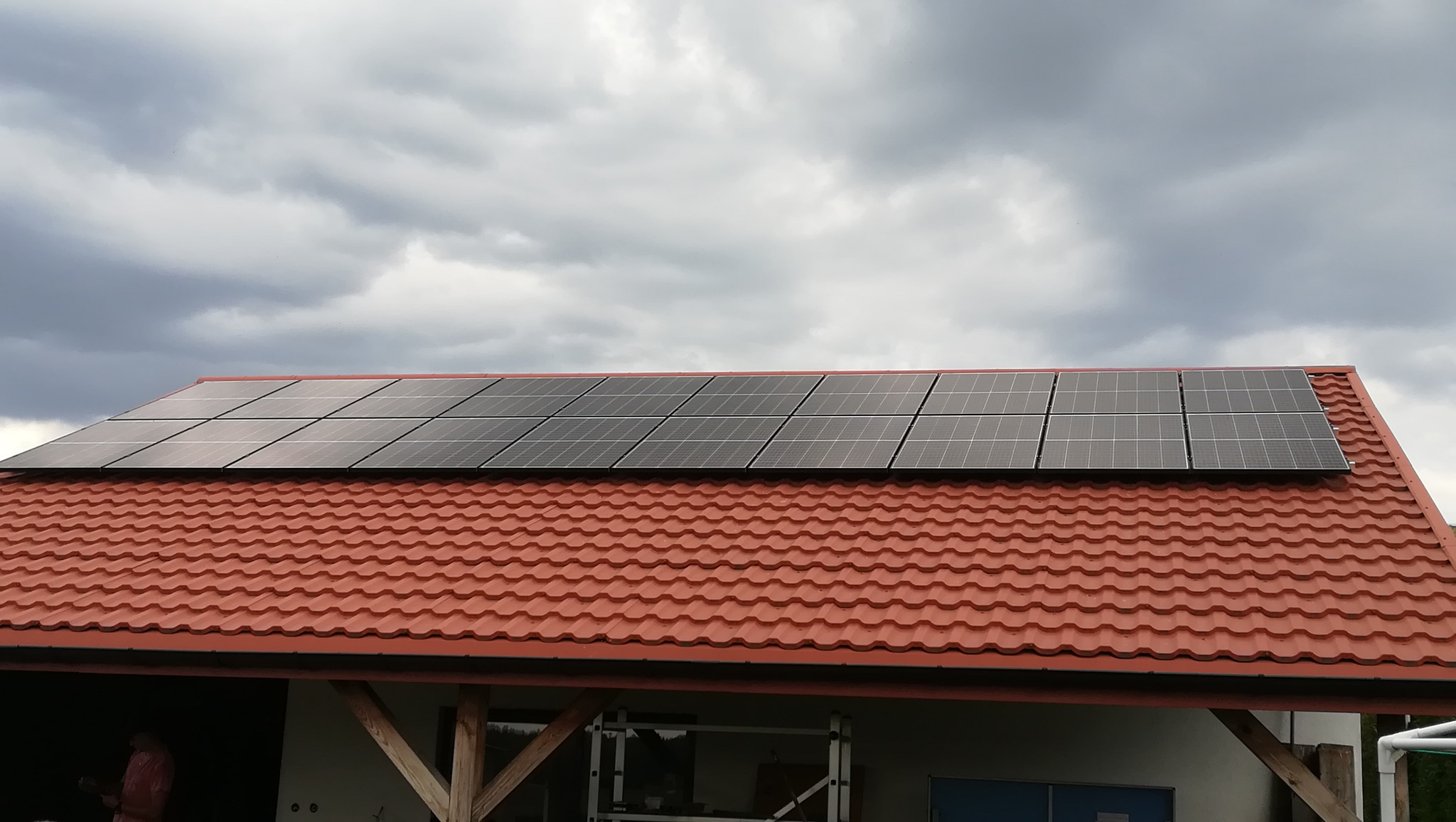 Panele słoneczne na dachu z czerwonej dachówki, widok na pochmurne niebo, częściowo widoczny budynek w tle, drabina rusztowania przy ścianie.