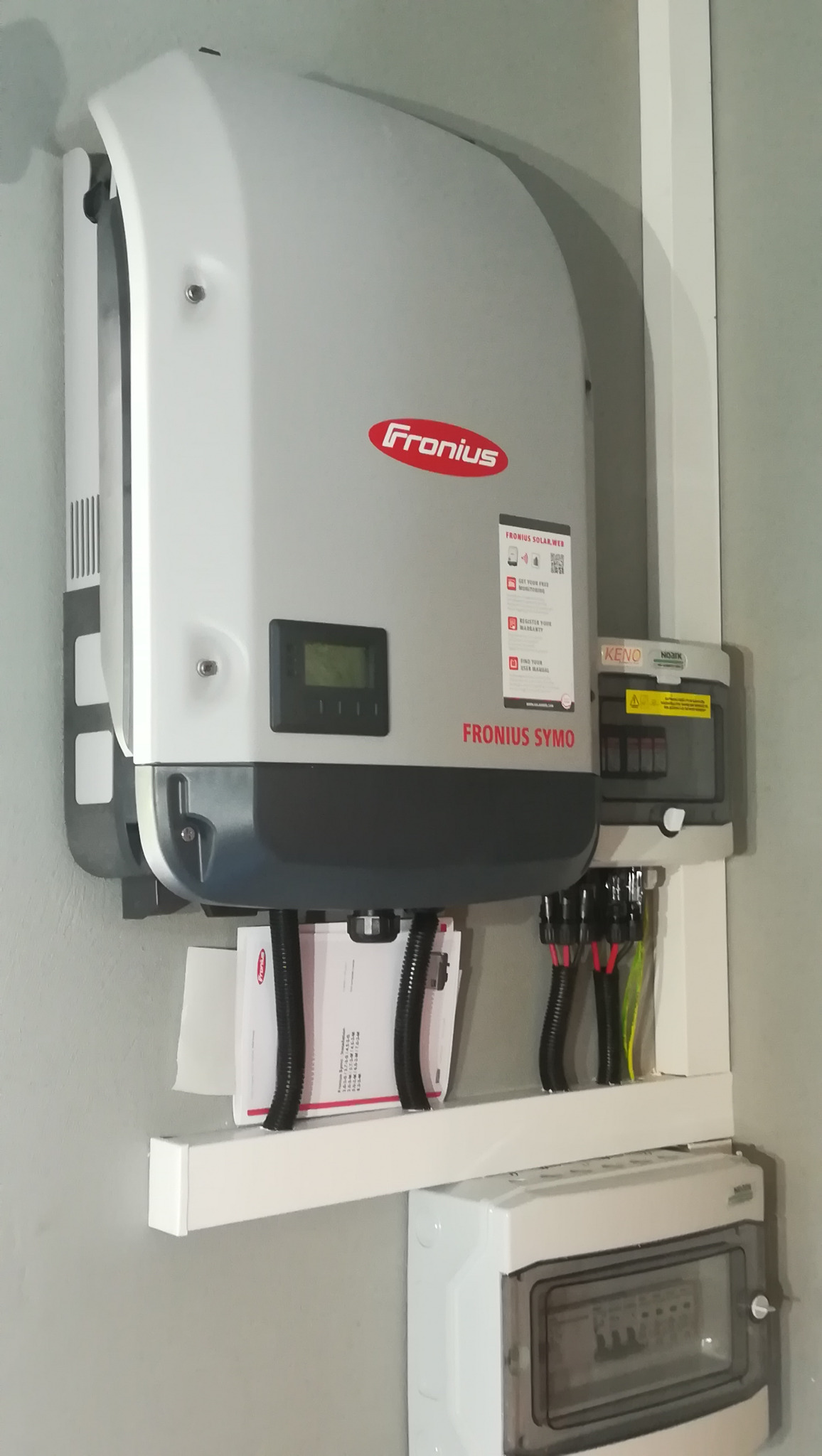Szary inwerter Fronius Symo zamontowany na ścianie, z widocznymi przewodami w peszlach i dokumentacją pod spodem, obok skrzynka zabezpieczeń Keno Noark.