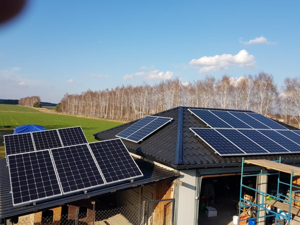 Panele słoneczne na dachu garażu z widokiem na pole i las w tle, instalacja w trakcie montażu, widoczne rusztowania.