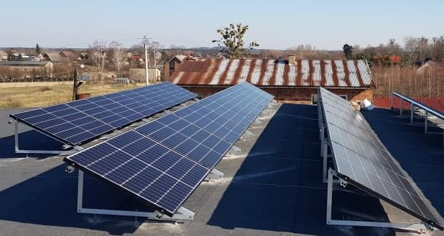 Instalacja paneli słonecznych na płaskim dachu z widokiem na zabudowania wiejskie i drzewa bez liści w tle, uchwycona w słoneczny dzień.