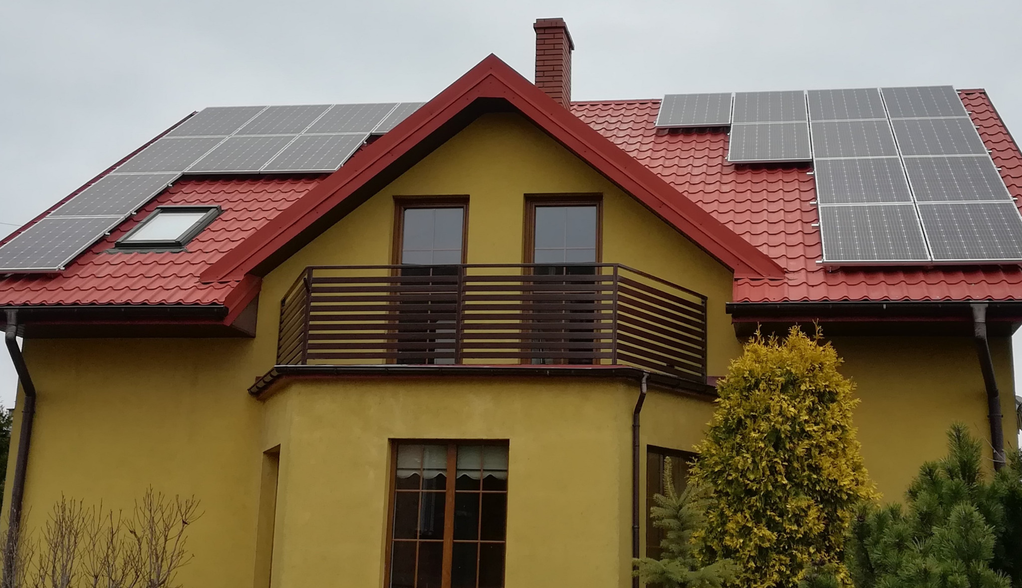Żółty dom jednorodzinny z czerwonym dachem, na którym zamontowano panele słoneczne w różnych konfiguracjach, widoczne okno dachowe i balkon z brązową balustradą.