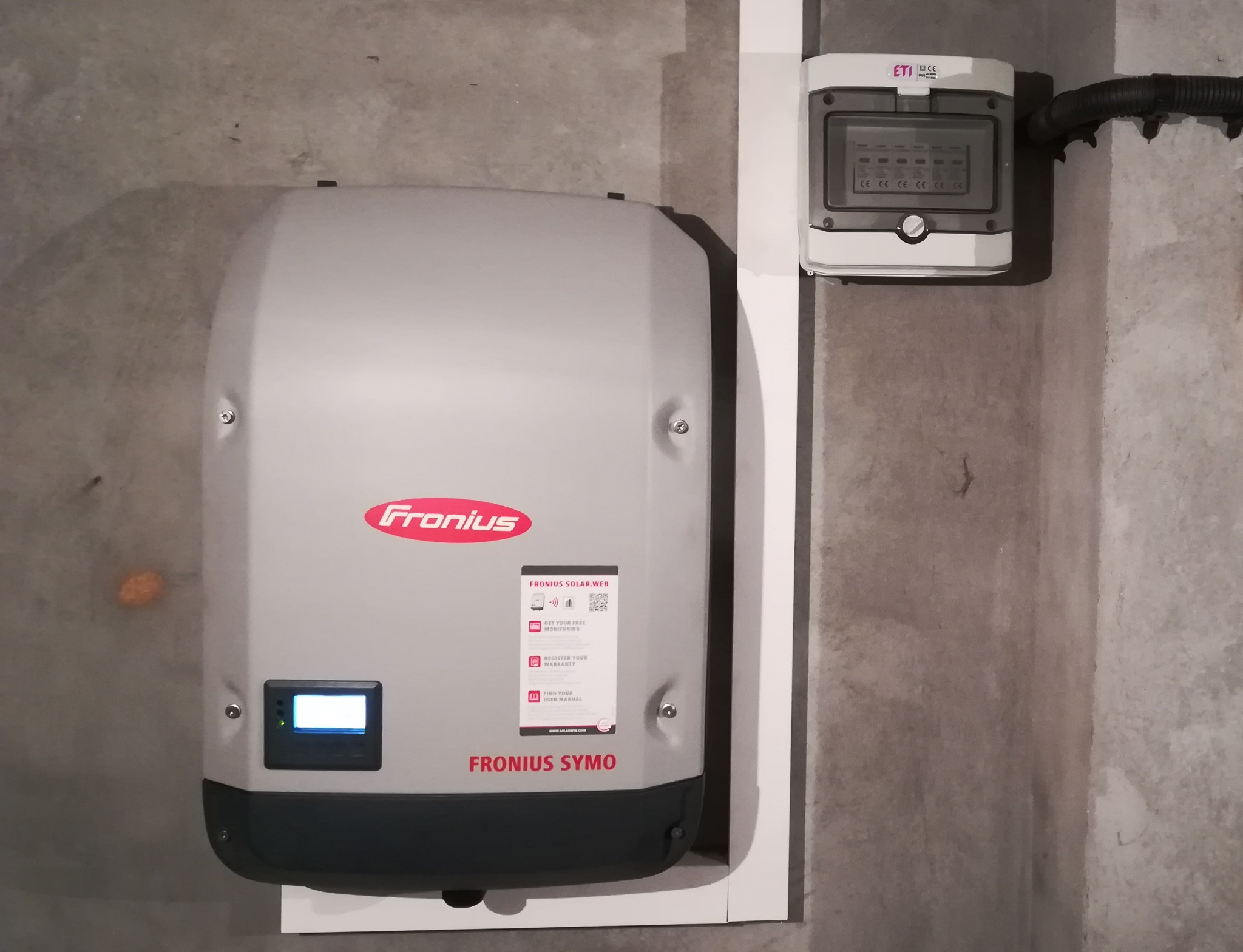 Szary falownik Fronius Symo zamontowany na ścianie z betonu architektonicznego, obok biała listwa i skrzynka elektryczna z widocznymi bezpiecznikami.