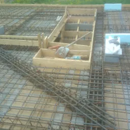 Zbrojenie płyty fundamentowej z drewnianym szalunkiem, widoczna łopata i cegły, styropian, siatka z prętów zbrojeniowych tworząca regularną strukturę.
