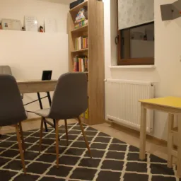 Jasne biuro z dwoma szarymi krzesłami, biurkiem, żółtą lampką, regałem z książkami, dywanem w romby i dziecięcym stolikiem z krzesełkiem, na ścianach dyplomy i dekoracje.