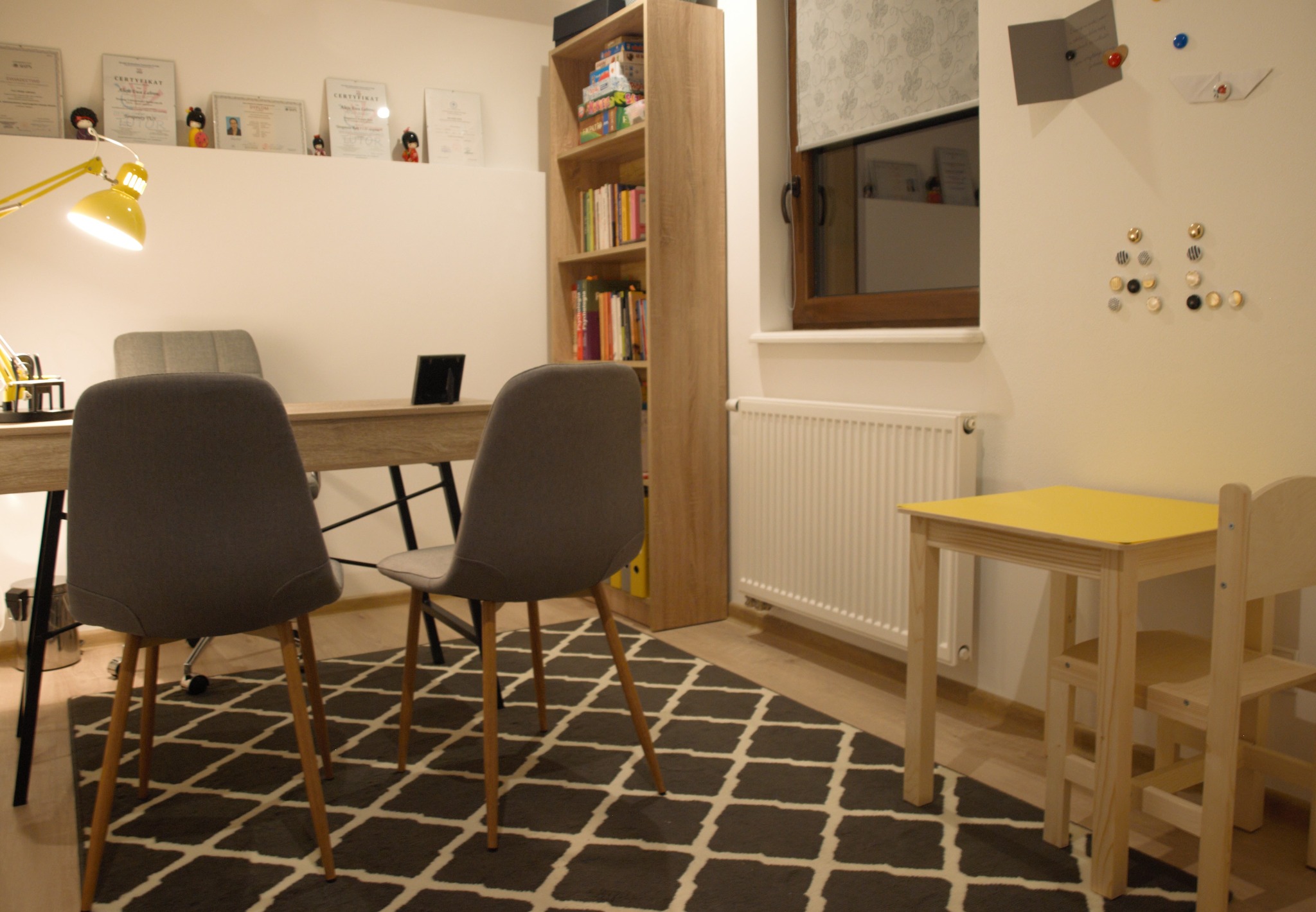Jasne biuro z dwoma szarymi krzesłami, biurkiem, żółtą lampką, regałem z książkami, dywanem w romby i dziecięcym stolikiem z krzesełkiem, na ścianach dyplomy i dekoracje.