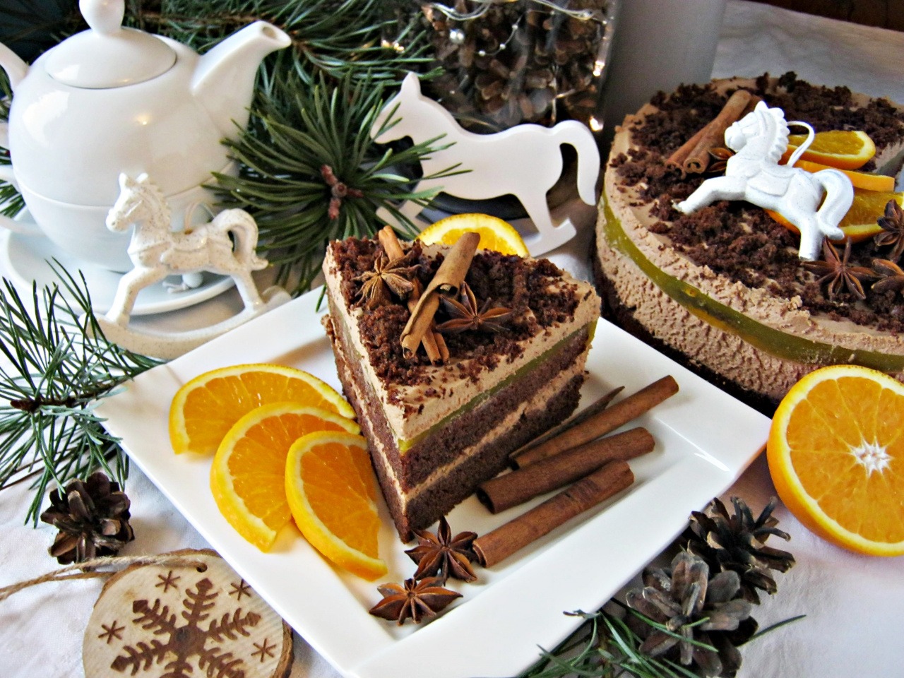 Kawałek tortu czekoladowego z kremem, pomarańczami, anyżem i laskami cynamonu na białym talerzu, w tle cały tort, biały imbryk i dekoracje świąteczne.