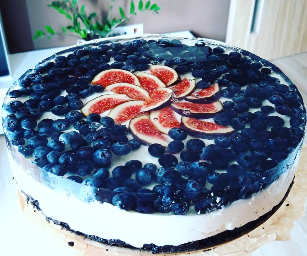 Tort z galaretką, borówkami i figami ułożonymi w kształt kwiatu, na tle zielonej rośliny doniczkowej.