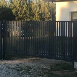 Nowoczesna, przesuwna brama wjazdowa z metalowymi sztachetami w kolorze grafitowym, widoczna z perspektywy podjazdu wysypanego żwirem, z domem i wysokimi tujami w tle.
