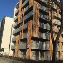 Nowoczesny budynek wielorodzinny z balkonami zabezpieczonymi szarą balustradą i pionowymi drewnianymi panelami dekoracyjnymi, widok z dołu na tle błękitnego nieba, z przodu drzewo bez liści.